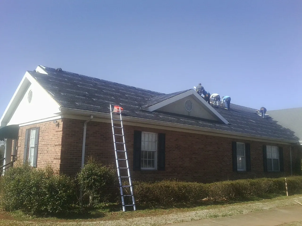 Crew tearing shingles off a brick ranch roof with a ladder set — Emergency Roof Repair work by our crew in New Richland