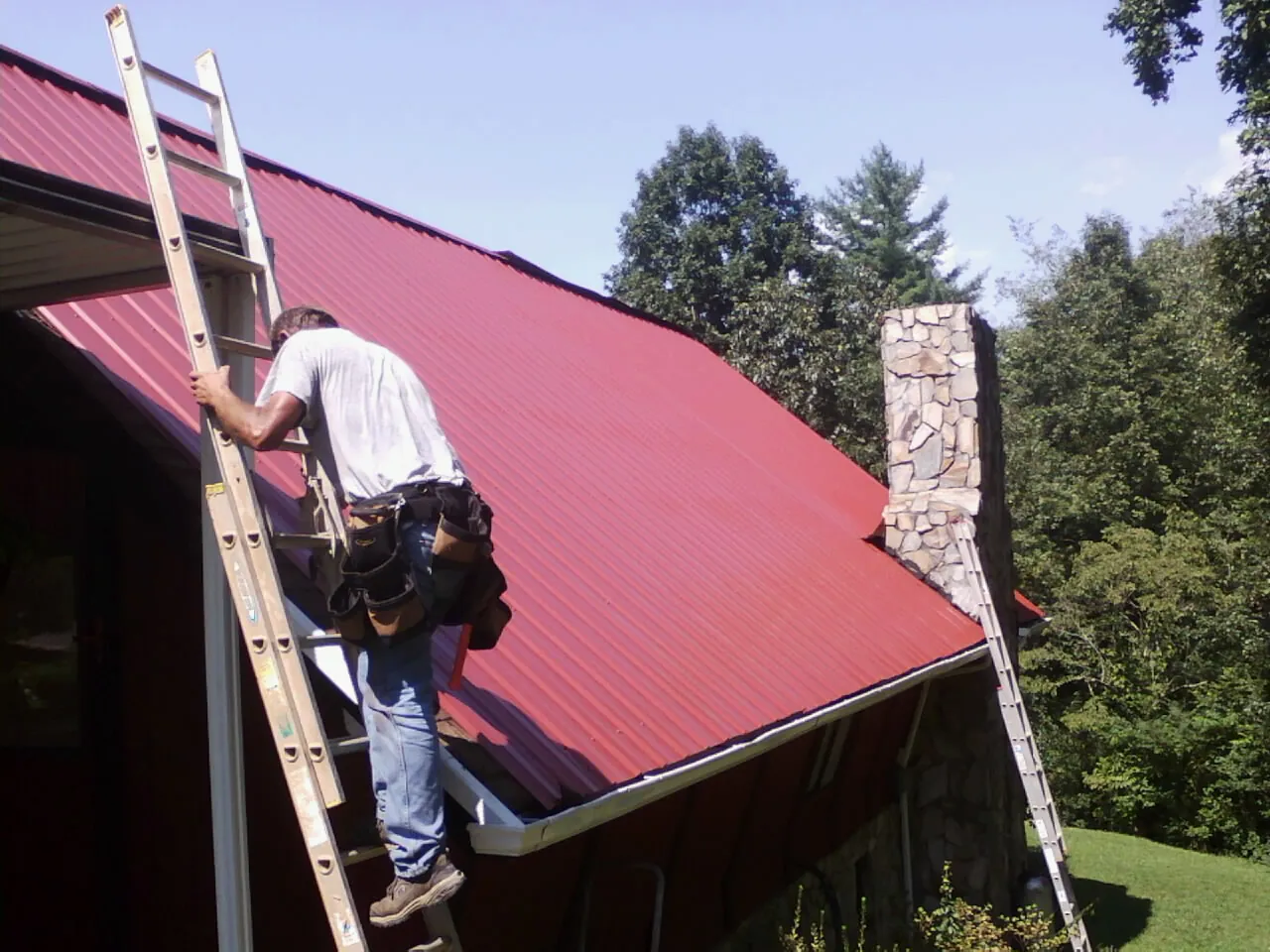 Roofer on a ladder beside a red metal roof and stone chimney — Soffit & Fascia Repair by Roofing Brothers in New Richland